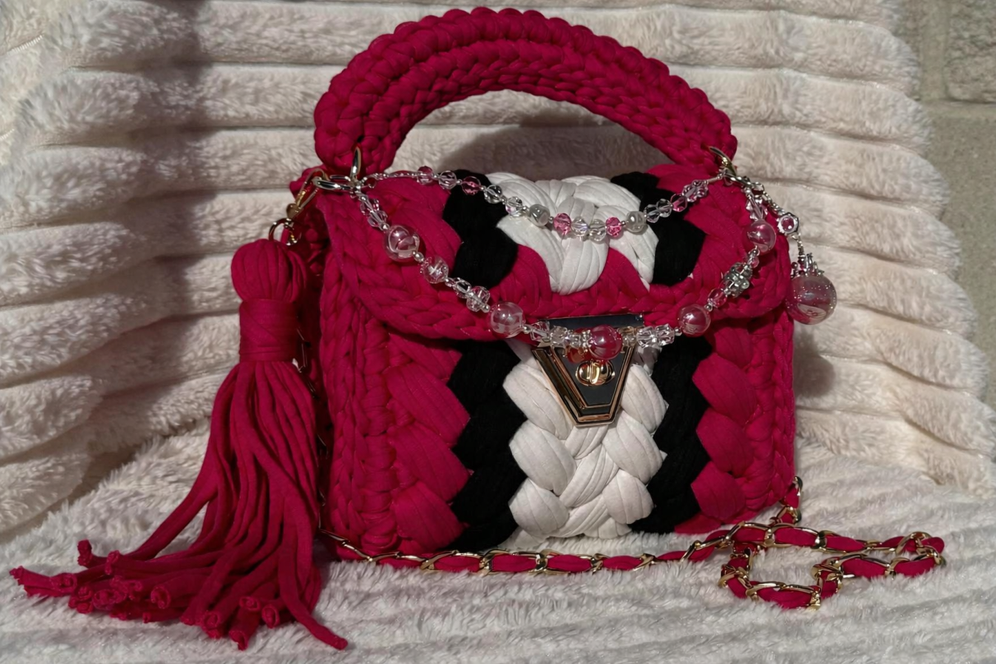 Front/Side View of the bag in full sunlight: Hot Pink, Black, and White Crochet woven handbag with Keychain and decorative elements sitting on a textured White surface