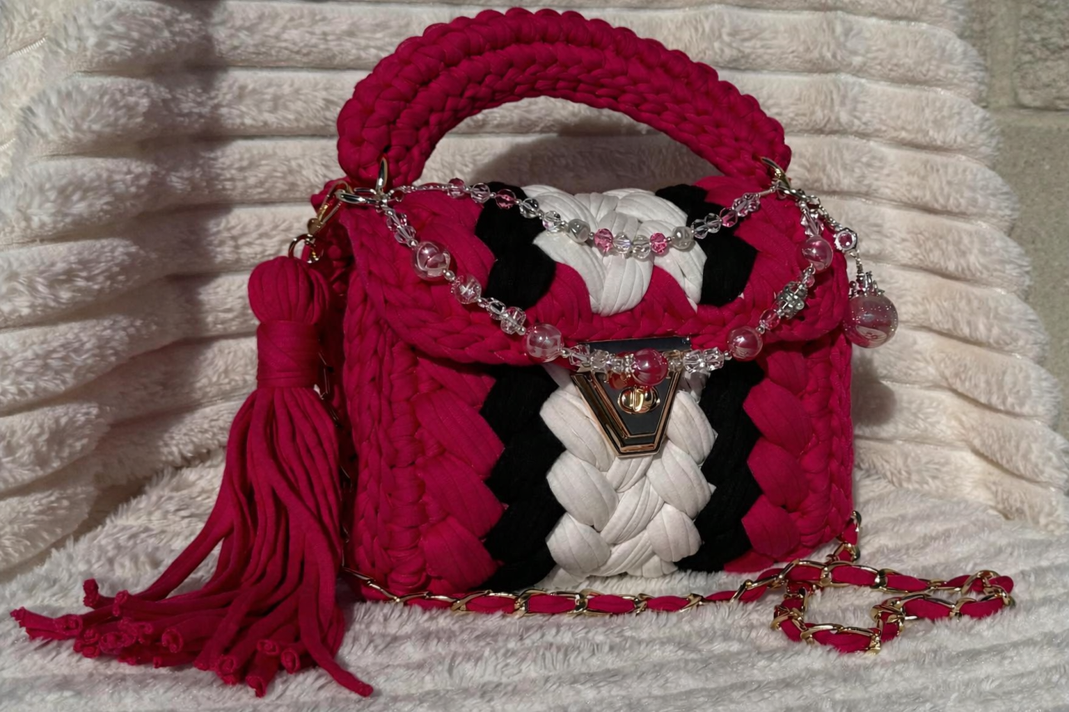 Front/Side View of the bag in full sunlight: Hot Pink, Black, and White Crochet woven handbag with Keychain and decorative elements sitting on a textured White surface