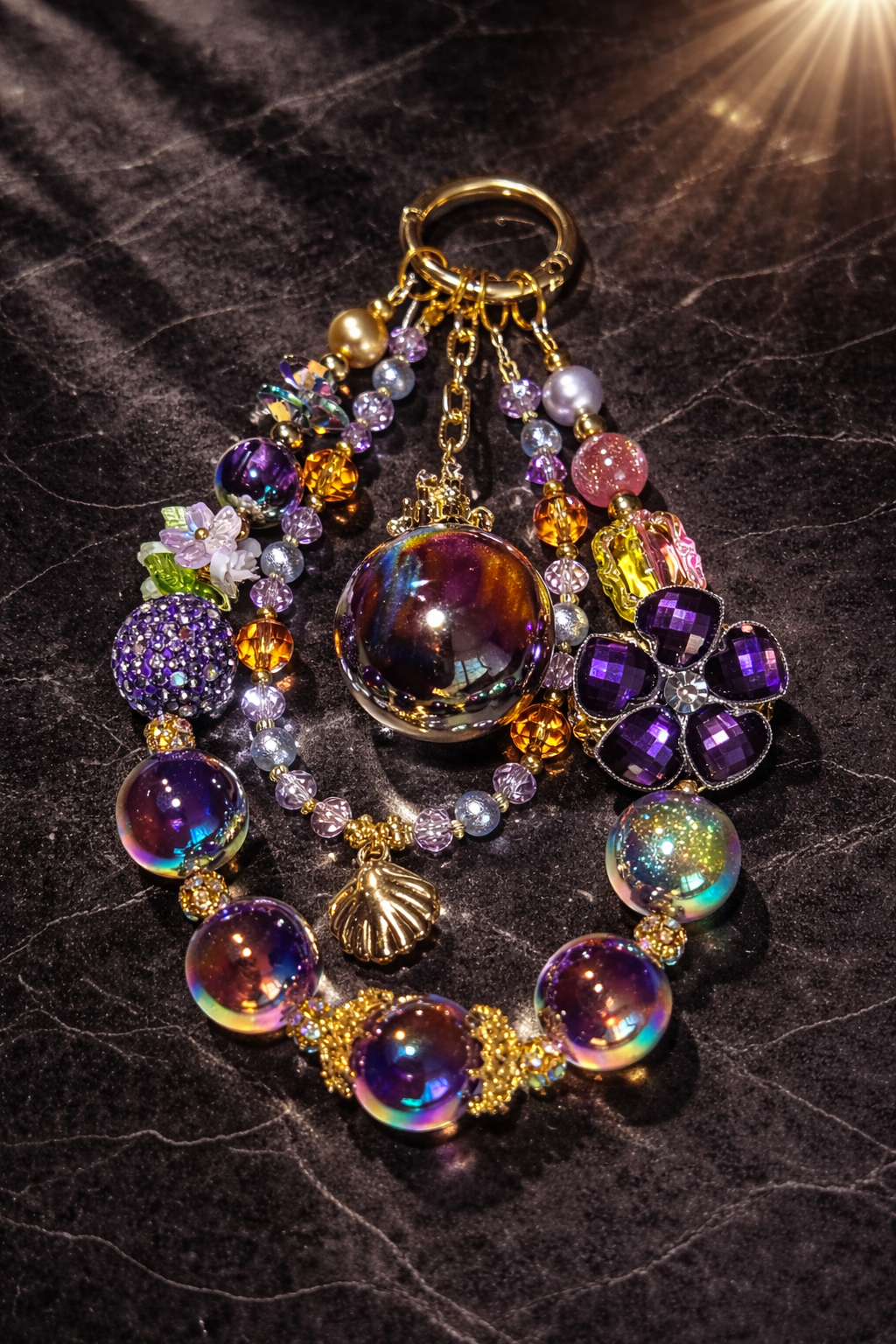 Purple decorative keychain with purple-iridescent beads, glittery gemstones, and a seashell pendant on a dark marble surface. 