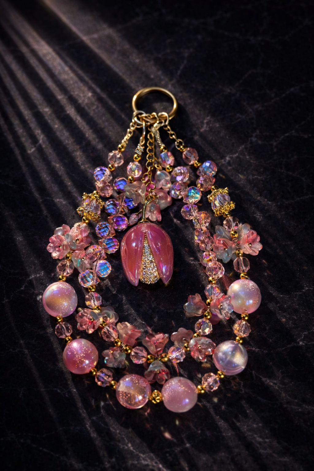 Large decorative gold and pink keychain with shimmering pink-pearl beads, purple-blue sparkling crystal beads, and pink flower accents on a dark textured marble background. 
