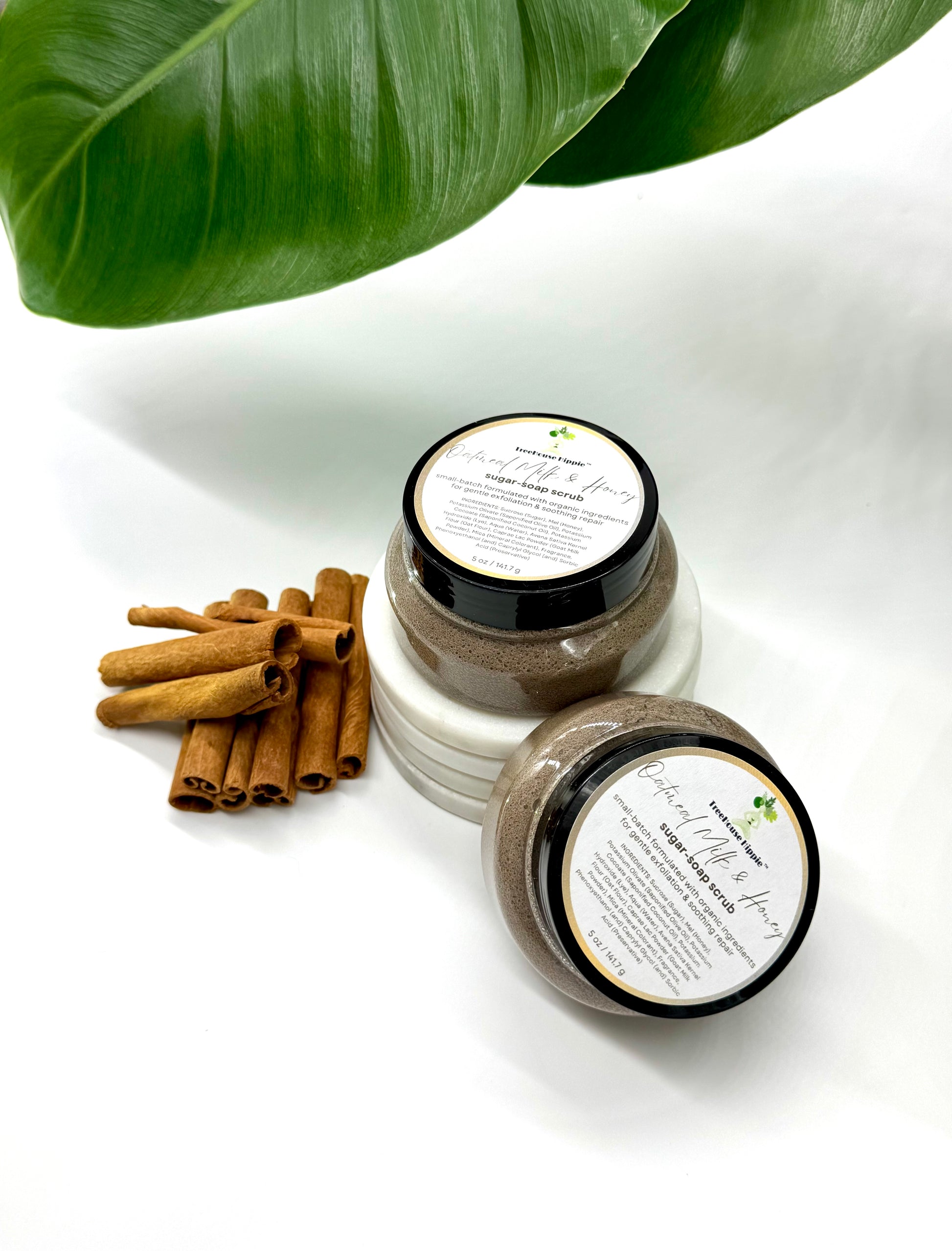 Two of TreeHouse Hippie's 'Oatmeal, Milk, & Honey' scrubs sitting beside cinnamon sticks on a white background with leaf accents.