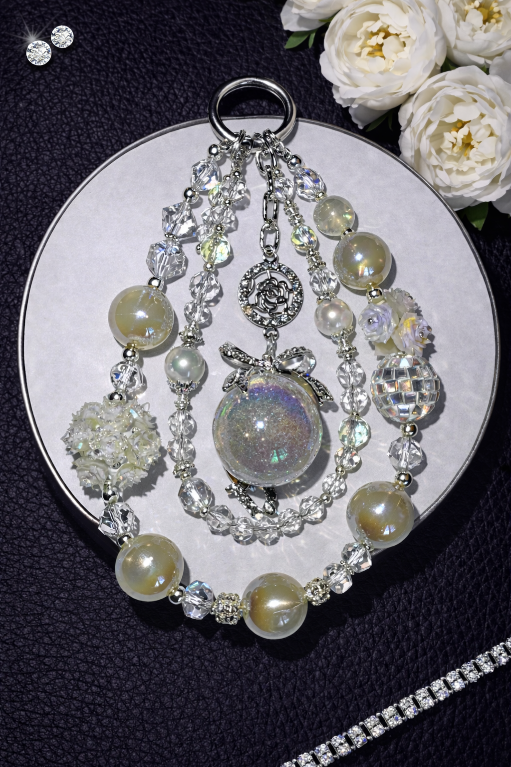 Decorative silver keychain with white pearl beads and white floral accents on a dark leather surface with white flowers in the top right corner and a silver chain in the bottom right corner. 