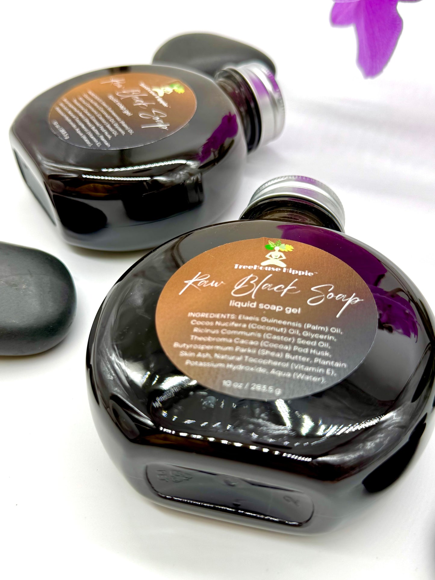 Two large bottles of TreeHouse Hippie's Raw Black Soap Gel with purple orchids and massage stones on a white background. 