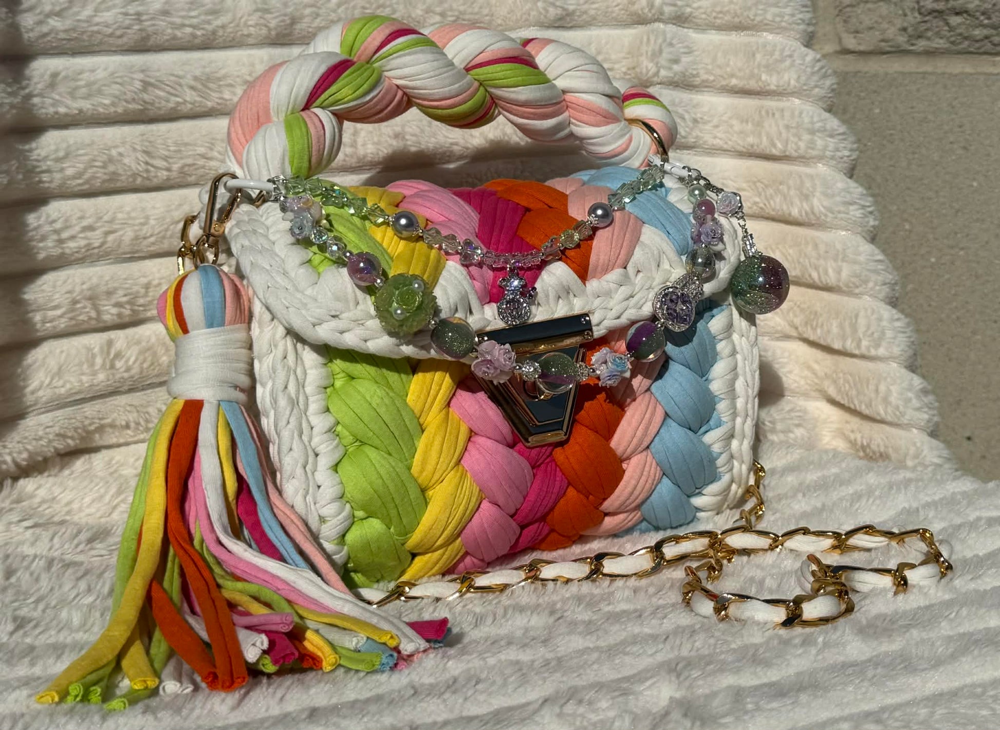 Side/Front view of bag in full sun: Colorful woven Crochet handbag with pastel rainbow pattern, tassel, and shimmering keychain sitting on a white, textured surface. Pastel Rainbow pattern includes: Lime Green, Orange, Yellow, Light Blue, Pink, and White. 