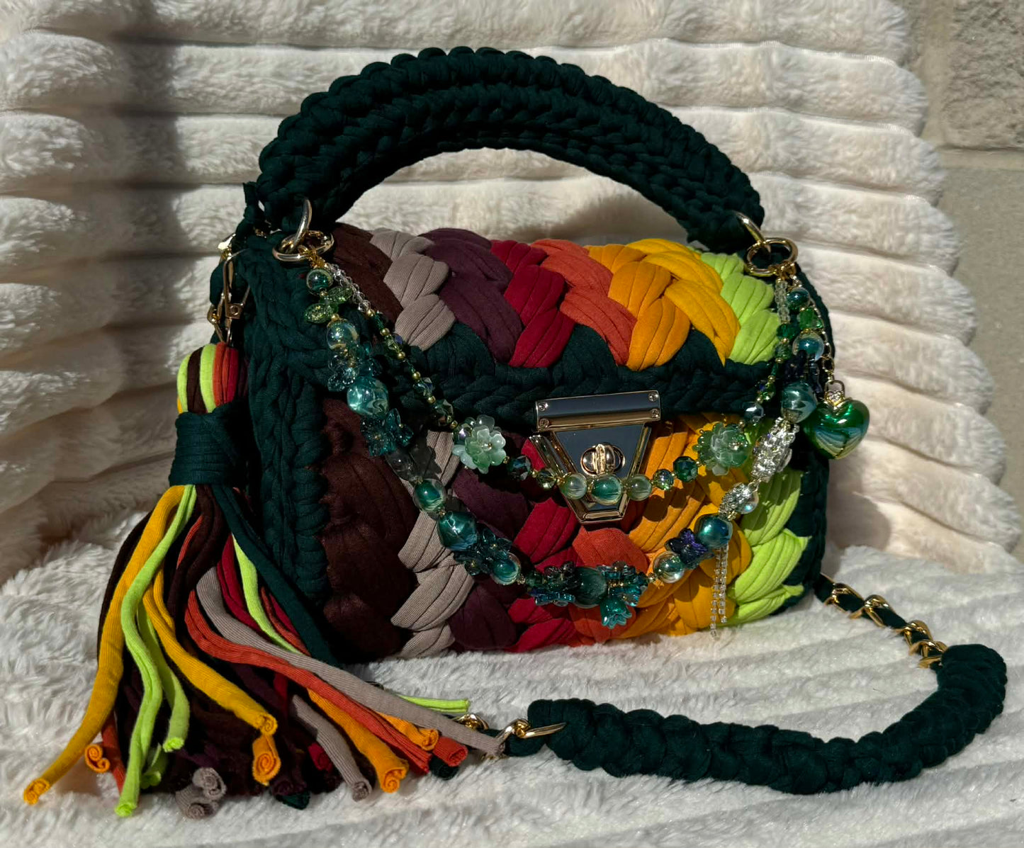 Side/Front view of bag in full sun: Colorful woven Crochet handbag (Forrest Green, Red, Burgundy, Lime Green, Orange, Yellow, and Gold) with tassel and beautiful shimmering keychain sitting on a white, textured surface. 