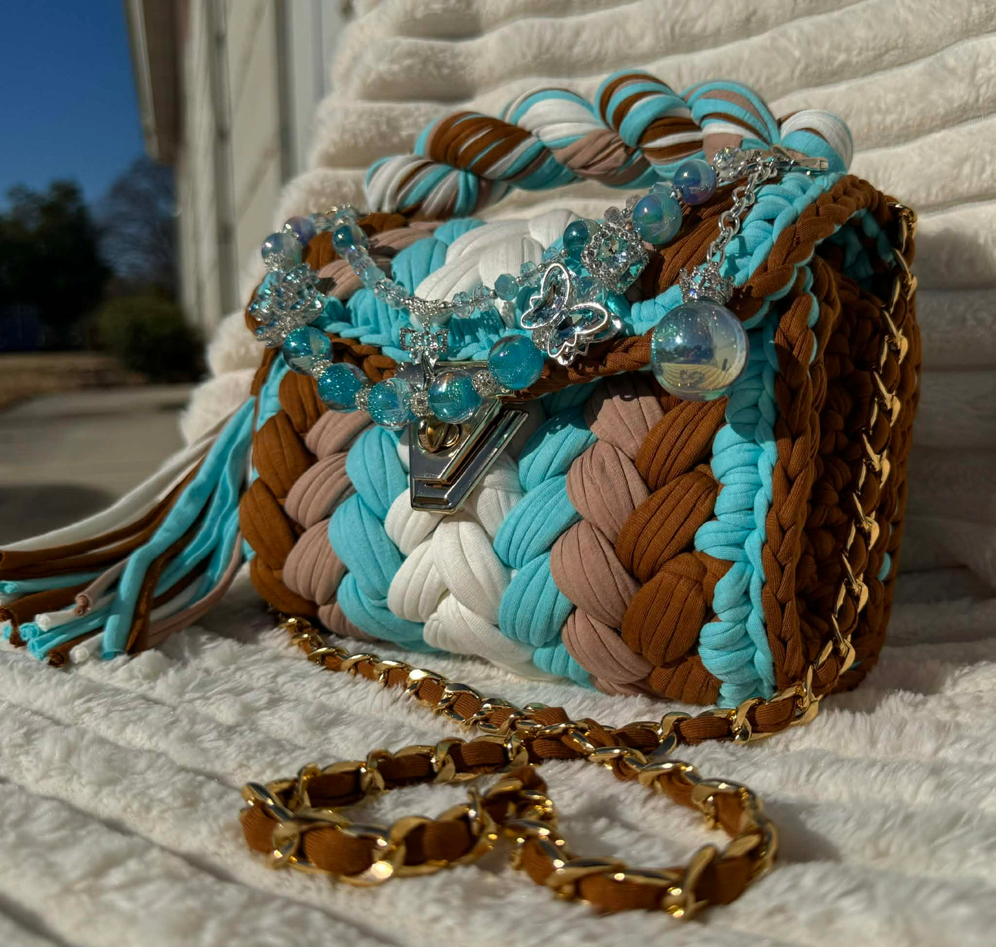 Side view of bag in full sun: Colorful woven Crochet handbag (White, Brown, Turquoise, & Milk Chocolate) with tassel and beautiful shimmering keychain sitting on a white, textured surface. 