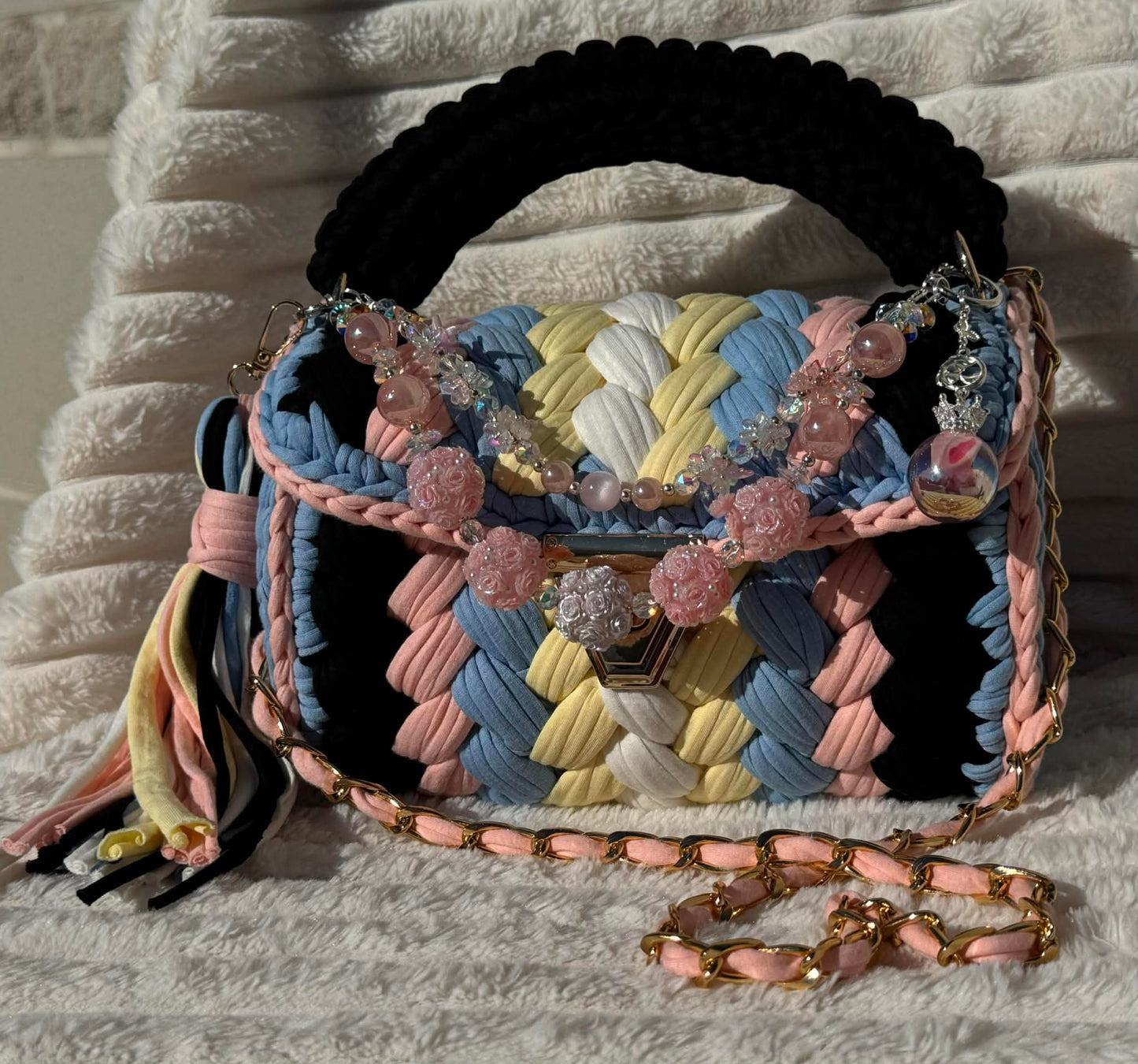 Front view in full sun: Multicolored woven Crochet handbag with decorative keychain and tassel sitting on a textured white background. Bag colors include: Black, Light Blue, Pink, Soft Yellow, and White. 