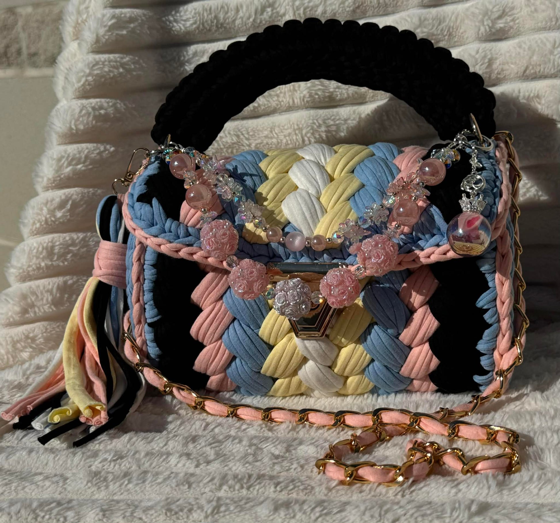 Front view in full sun: Multicolored woven Crochet handbag with decorative keychain and tassel sitting on a textured white background. Bag colors include: Black, Light Blue, Pink, Soft Yellow, and White. 
