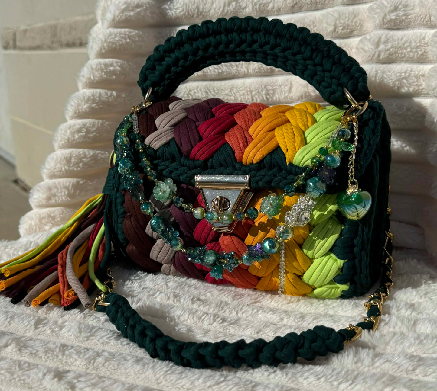 Front view of bag in full sun: Colorful woven Crochet handbag (Forrest Green, Red, Burgundy, Lime Green, Orange, Yellow, and Gold) with tassel and beautiful shimmering keychain sitting on a white, textured surface. 