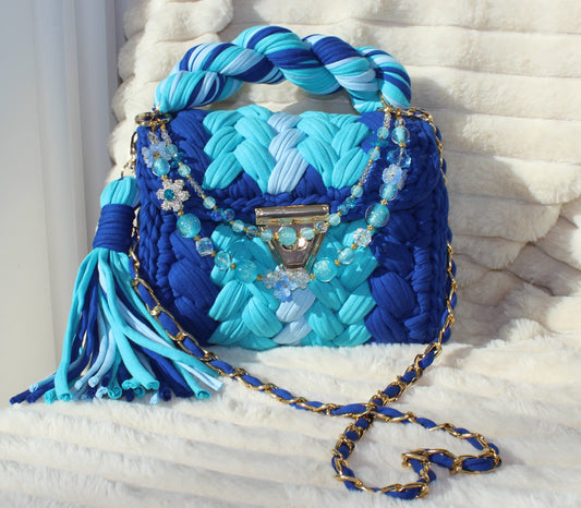 Front view of bag in natural lighting: Woven Crochet handbag (Royal Blue, Light Blue, and Ice Blue)  with tassel and beautiful shimmering keychain sitting on a white, textured surface. 